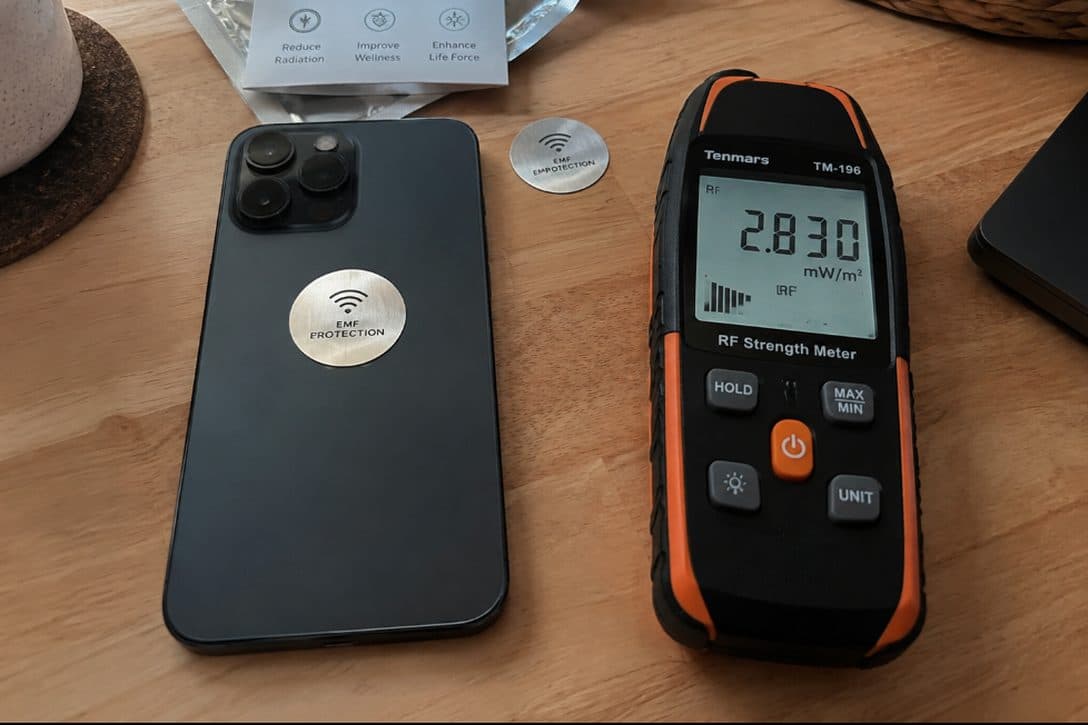 A phone with an EMF sticker attached beside a handheld RF meter reading 28.30.